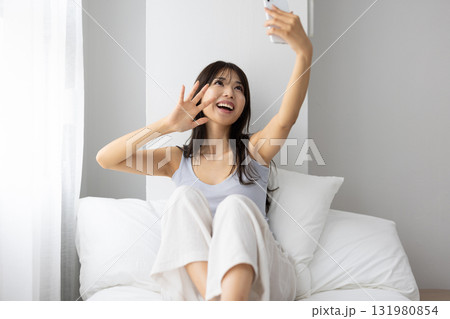 ベッドの上で窓からの朝日を浴びながらスマートフォンでビデオ通話する若い女性若い女性 ベッドの上で窓からの朝日を浴びながらスマートフォンでビデオ通話する若い女性若い女性 131980854