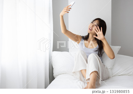 ベッドの上で窓からの朝日を浴びながらスマートフォンでビデオ通話する若い女性若い女性 ベッドの上で窓からの朝日を浴びながらスマートフォンでビデオ通話する若い女性若い女性 131980855