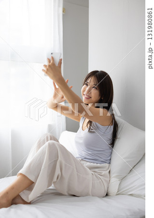 ベッドの上で窓からの朝日を浴びながらスマートフォンでビデオ通話する若い女性若い女性 ベッドの上で窓からの朝日を浴びながらスマートフォンでビデオ通話する若い女性若い女性 131980861