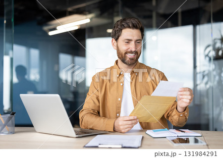 Happy bearded man sitting at a modern office desk opening an envelope and reading a document, celebrating success, and enjoying positive business communication Happy bearded man sitting at a modern office desk opening an envelope and reading a document, celebrating success, and enjoying positive business communication 131984291