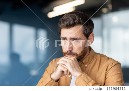 Man sits with hands clasped under his chin, pensive and worried in a modern office, contemplating business challenges, strategy and solutions amid stress and responsibility 131984311