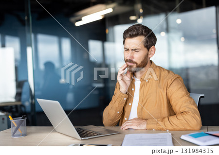 Young man working on laptop in a modern office, experiencing severe toothache and jaw pain, touching his cheek with hand, showing discomfort and dental problem Young man working on laptop in a modern office, experiencing severe toothache and jaw pain, touching his cheek with hand, showing discomfort and dental problem 131984315