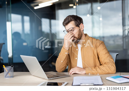 Businessman feeling overwhelmed and tired, rubbing his eyes from eye strain and headache while working on a laptop at an modern office desk, symbolizing work-related stress and fatigue Businessman feeling overwhelmed and tired, rubbing his eyes from eye strain and headache while working on a laptop at an modern office desk, symbolizing work-related stress and fatigue 131984317