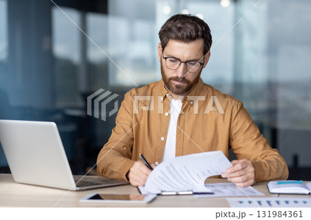 Confident focused businessman working on paperwork while sitting at a desk, reviewing a business contract and preparing to sign it in a modern office setup 131984361