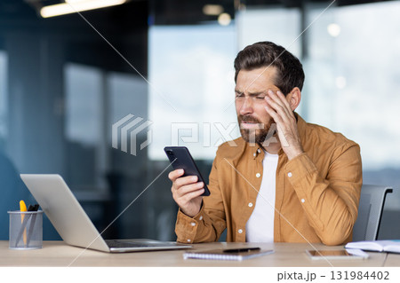 Stressed businessman receives bad news on his cellphone, clutching his forehead in a modern office, showing anxiety, headache and frustration over work or financial trouble 131984402