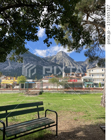 A bench stands under a tree in the park against the backdrop of mountains and houses A bench stands under a tree in the park against the backdrop of mountains and houses 131985327