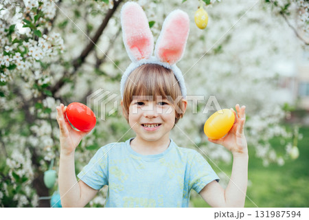 Easter Egg Hunt with Child Carrying Bright Plastic Eggs. Smiling child wearing bunny ears holding a wicker basket filled with colorful Easter eggs in a blooming spring garden 131987594
