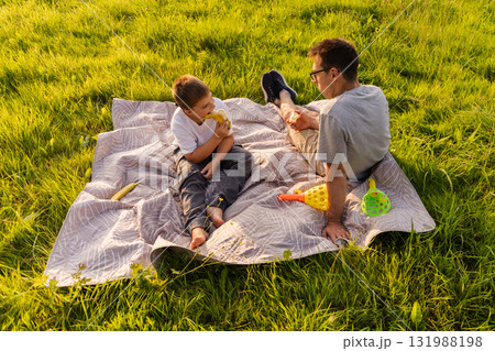 A father and his young son relax on a blanket in a lush green park. They share snacks and play with colorful toys, enjoying a sunny afternoon. Laughter fills the air as they bond A father and his young son relax on a blanket in a lush green park. They share snacks and play with colorful toys, enjoying a sunny afternoon. Laughter fills the air as they bond 131988198