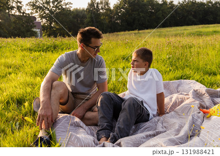 A father sits on a blanket in a grassy field, facing his son, who looks angry. They are spending time together in nature during late afternoon, surrounded by trees and open space 131988421