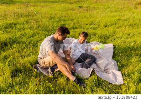 A father and son sit on a blanket in a lush green field, laughing and playing together. The sun shines bright in the afternoon sky as they cherish these precious moments A father and son sit on a blanket in a lush green field, laughing and playing together. The sun shines bright in the afternoon sky as they cherish these precious moments 131988423
