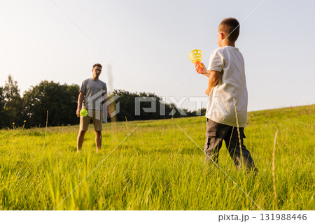 A father and his young son are having fun together in a lush green field. They are playing catch with colorful balls as the sun shines brightly on a warm afternoon 131988446