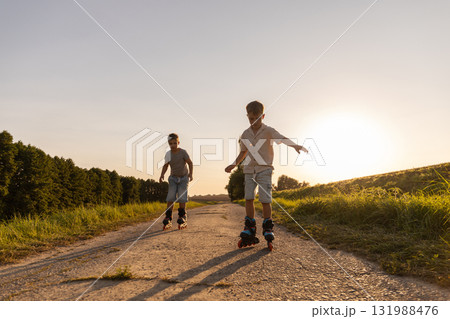 Two friends are having fun inline skating down a dirt path surrounded by trees and grass. They are actively enjoying their free time in the warm evening light 131988476