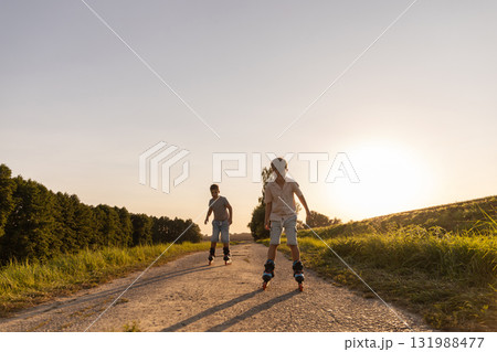 Two friends are having fun inline skating down a dirt path surrounded by greenery under a clear blue sky during late afternoon sunshine 131988477