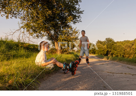 Two friends are inline skating on a warm afternoon. One child sits on the grass, while the other skates down the path, surrounded by trees and nature 131988501