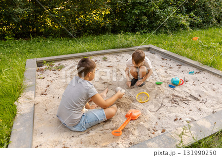 Two kids are playing in a sand pit surrounded by greenery. They are using colorful toys to build structures and enjoy each other's company on a sunny day Two kids are playing in a sand pit surrounded by greenery. They are using colorful toys to build structures and enjoy each other's company on a sunny day 131990250