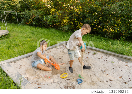 Two children are happily playing in a sandbox, digging with colorful shovels. Lush green grass and trees create a natural backdrop in a warm afternoon setting, enhancing their fun Two children are happily playing in a sandbox, digging with colorful shovels. Lush green grass and trees create a natural backdrop in a warm afternoon setting, enhancing their fun 131990252