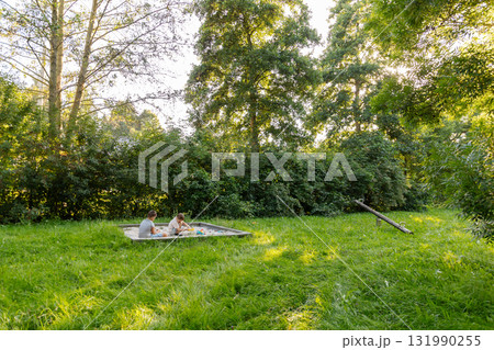 Two kids are actively engaged in play in a lush green area filled with trees. The setting is serene, showcasing nature's beauty during a sunny afternoon Two kids are actively engaged in play in a lush green area filled with trees. The setting is serene, showcasing nature's beauty during a sunny afternoon 131990255