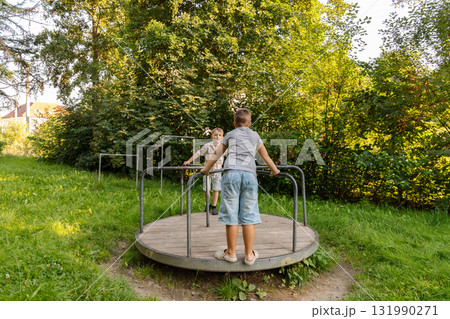 Two children are having fun on a merry-go-round in a lush green park during a sunny afternoon. They are smiling and playing together, enjoying the great outdoors 131990271
