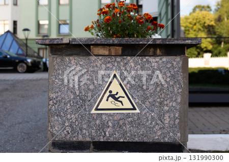 Cautionary slip hazard sign, affixed to a stone pillar with vibrant flowers on top. Reminder of safety in public spaces, promoting awareness of potential risks Cautionary slip hazard sign, affixed to a stone pillar with vibrant flowers on top. Reminder of safety in public spaces, promoting awareness of potential risks 131990300