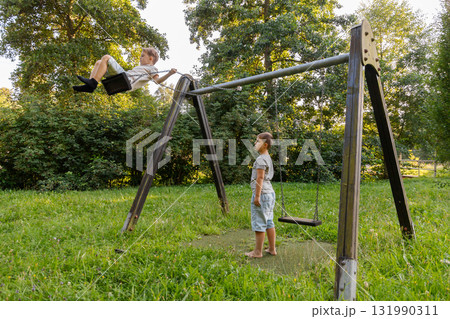 Two kids are playing joyfully in a green park. One child swings high while the other watches with excitement. Bright sunshine filters through the trees, creating a lively scene Two kids are playing joyfully in a green park. One child swings high while the other watches with excitement. Bright sunshine filters through the trees, creating a lively scene 131990311