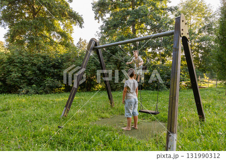 Two kids are having fun in a park on a swing set. One child swings happily while the other watches and enjoys the warm, sunny day surrounded by trees and grass Two kids are having fun in a park on a swing set. One child swings happily while the other watches and enjoys the warm, sunny day surrounded by trees and grass 131990312