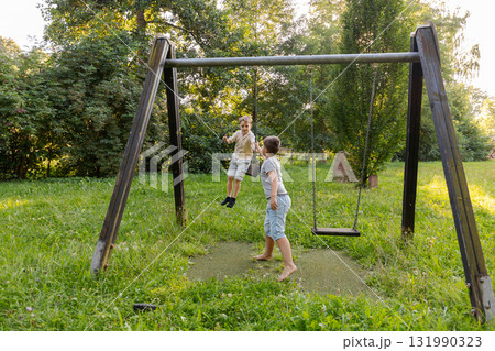 Two children are happily playing on swings in a vibrant park. The scene is filled with lush green grass and trees, radiating a joyful summer vibe Two children are happily playing on swings in a vibrant park. The scene is filled with lush green grass and trees, radiating a joyful summer vibe 131990323