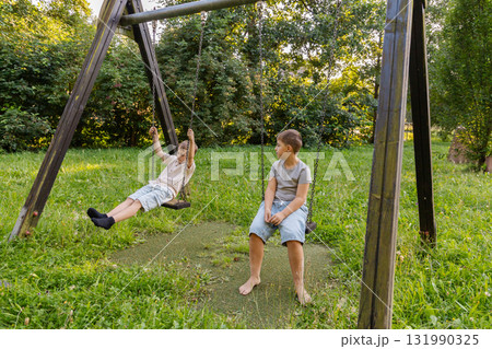 Two kids are playing joyfully on a swing set in a lush green area filled with trees. One child swings while the other watches, enjoying the beautiful sunny day Two kids are playing joyfully on a swing set in a lush green area filled with trees. One child swings while the other watches, enjoying the beautiful sunny day 131990325