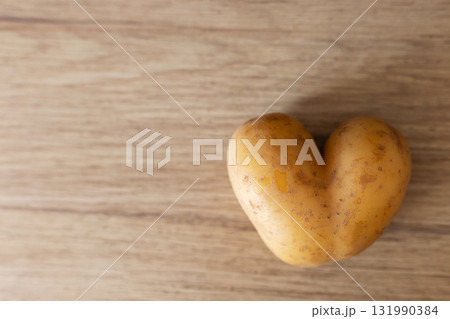 A unique heart-shaped potato rests on a smooth wooden table, showcasing its distinct shape and texture. The simple setting adds warmth, making it an intriguing sight for food lovers 131990384
