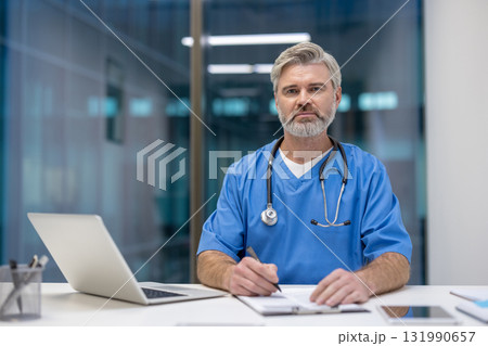 Male doctor in scrubs with stethoscope at a desk, writing on a clipboard while looking at the camera, conveying experience, focus and trustworthy patient care in a modern clinic 131990657