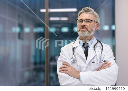Mature male doctor in white coat with stethoscope and glasses, arms crossed, looking thoughtfully into the distance in a bright modern clinic, conveying trust and expertise 131990660