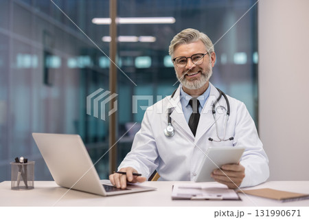 Mature doctor man smiling, wearing a stethoscope and white coat, working in his office with a laptop and digital tablet, showing innovation and modern healthcare 131990671