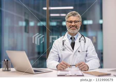 Experienced male doctor in white coat with stethoscope sits at clinic desk with laptop and documents, smiling confidently at the camera conveying trustworthy medical care 131990679