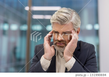 Senior businessman with gray hair and a beard, feeling intense pain and headache, holding temples with both hands, showing expressions of stress, frustration, and worry Senior businessman with gray hair and a beard, feeling intense pain and headache, holding temples with both hands, showing expressions of stress, frustration, and worry 131990694