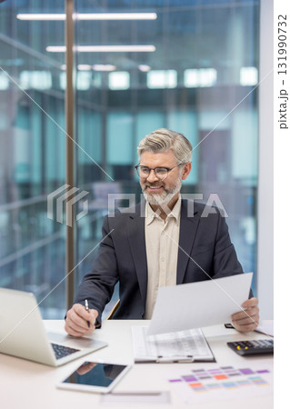 Businessman with gray hair and beard smiling while reviewing documents and using a laptop and tablet at his desk in a contemporary office setting with large window 131990732