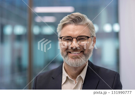 A portrait of a smiling mature businessman wearing glasses, showcasing confidence and success. The office background adds a professional atmosphere. 131990777