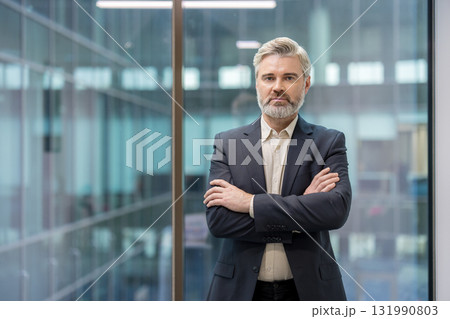Mature businessman with gray hair and beard in a dark suit standing confidently with crossed arms. Looking at the camera against a modern office background. Symbolizing leadership and professionalism Mature businessman with gray hair and beard in a dark suit standing confidently with crossed arms. Looking at the camera against a modern office background. Symbolizing leadership and professionalism 131990803