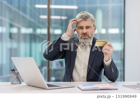Confused mature businessman scratching his head while holding a credit card, experiencing payment problems or financial debt issues during online shopping at his modern office desk 131990849