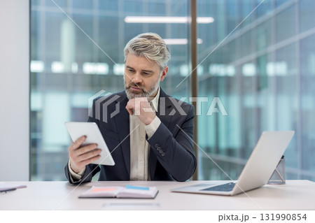 Experienced businessman in a suit intently reviewing data on a digital tablet while working at his desk in a contemporary office building with large windows 131990854