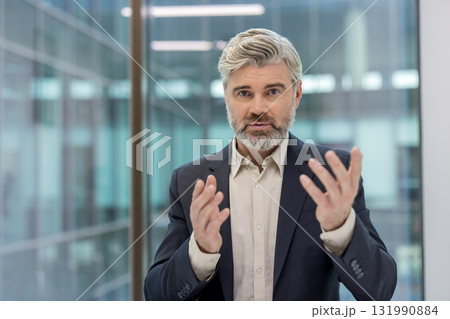 Senior businessman with a beard and grey hair speaking and gesturing with hands, sharing expertise and presenting ideas during a corporate conference in a modern office 131990884