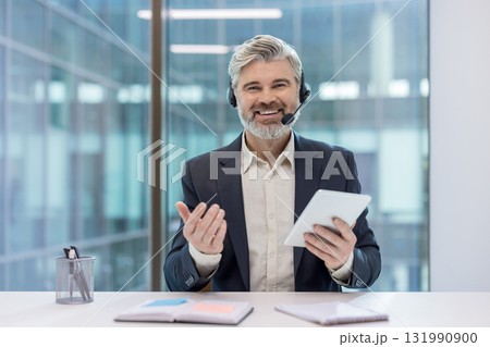 Mature man with a headset smiling and holding a digital tablet, representing concepts of online consultation, customer support, and virtual communication from a modern office environment 131990900