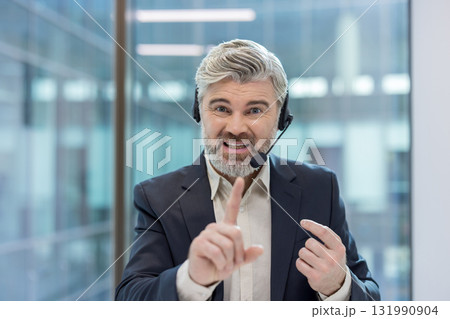 Mature man with a beard and headset talking, actively communicating with customers, providing support, and gesticulating with his finger in a modern office environment 131990904