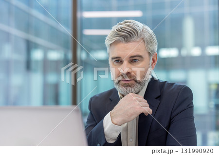 Mature businessman having a pensive expression, strategizing in a modern office environment while looking at a laptop screen, conveying deep thought and decision-making for business success 131990920