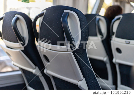 Passengers travel inside a bus, viewing the backs of comfortable seats with handles. Sunlight streams through the windows, illuminating the space and creating a welcoming atmosphere 131991239