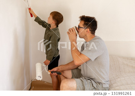 A father and son are working together to paint the bedroom walls white. The man is sitting and sipping a drink while the boy uses a roller brush to help with the task 131991261
