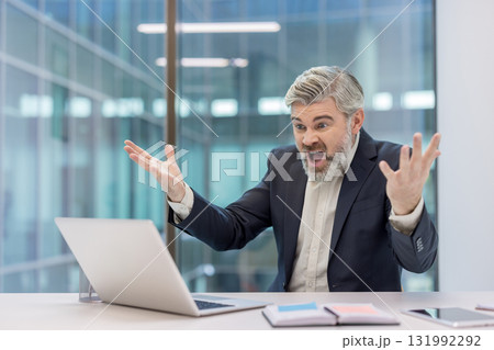 Agitated middle-aged businessman experiencing extreme stress and anger at his desk, reacting to a problem with his laptop in a modern office environment 131992292