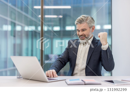Mature businessman celebrates a win at his modern office desk, smiling and raising a triumphant fist while looking at his laptop after a successful deal or breakthrough Mature businessman celebrates a win at his modern office desk, smiling and raising a triumphant fist while looking at his laptop after a successful deal or breakthrough 131992299