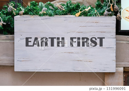 An Earth First sign, in dark letters on distressed white wood, is decorated with garland and pinecones. It hangs outdoors, perhaps near a shop during the daytime An Earth First sign, in dark letters on distressed white wood, is decorated with garland and pinecones. It hangs outdoors, perhaps near a shop during the daytime 131996091