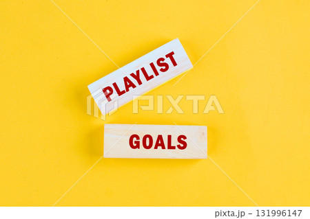 On a bright yellow background, two wooden blocks are placed showing the words PLAYLIST and GOALS, suggesting the act of creating musical experiences with intentional song choices 131996147