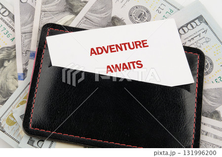 A black leather wallet containing a card that reads ADVENTURE AWAITS rests on a pile of United States one hundred dollar bills, signaling a planned journey A black leather wallet containing a card that reads ADVENTURE AWAITS rests on a pile of United States one hundred dollar bills, signaling a planned journey 131996200