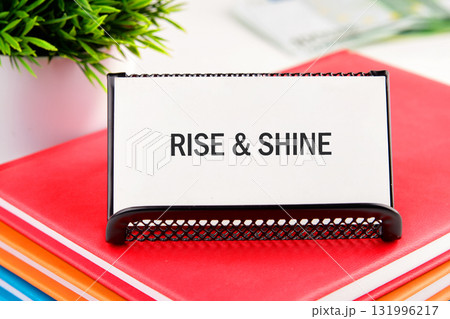 A black sign holder displays RISE SHINE on a white card. It sits atop a stack of vibrant books with a small potted plant in the background A black sign holder displays RISE SHINE on a white card. It sits atop a stack of vibrant books with a small potted plant in the background 131996217